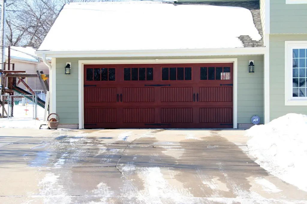 Dark red garage door with small windows across the top, in a pale green home with snow on the yard and on the roof.